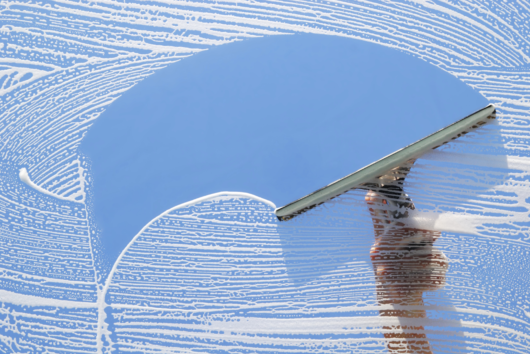 A squeegee clearing soapy window glass against a blue sky